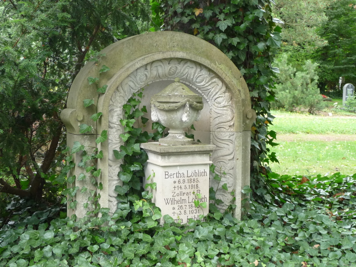 Altes Grabmal von 1918 aus Sandstein, mit Rundbogenabschluss. Vor dem Stein eine Säule mit Urnenaufsatz.