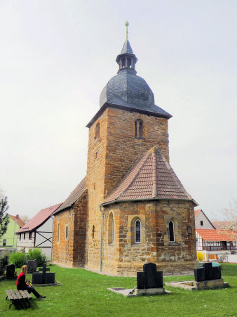 Blick zur Kirche über den Friedhof. Die Kirche aus Sandstein und mit Ziegeln gedeckt. Am Kirchturm schließt sich noch ein kleiner Kirchenteil an. Die Kuppel mit Schiefer des Turmes ist achteckig und mündet in einem Glockentürmchen.