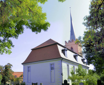 Hinter Gräbern auf dem Friedhof steht die hell angestrichen Kirche. Hohe Fenster prägen die Fassade, das mit Ziegeln gedeckte Dach hat drei Gauben. Dahinter ist der mit Zinnen besetzte Turm und die Kirchturmspitze zu erkennen.