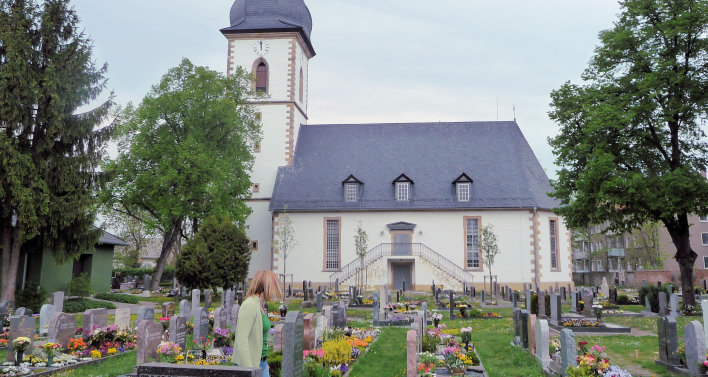 Blick über den Friedhof zur Längsseite der hell gestrichenen Kirche. Die Fensterrahmen und Gebäudekanten sind farbig abgesetzt. In der Mitte des Kirchenschiff befinden sich zwei übereinander liegende Eingänge. Der obere wird durch eine von links und rechts kommende Treppe erreicht. dach und Turm sind mit Schiefer eingedeckt. Am Kirchturm befindet sich über dem obersten Fenster eine Kirchturmuhr. 