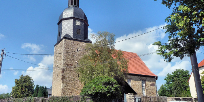 Blick von der Straße zur Kirche. Der Kirchhof ist mit einer Natursteinmauer eingefasst, auf der ein Holzzaun steht, teilweise mit Grün überwuchert. Die Kirche aus Natursteinen und einem roten Ziegeldach hat an der linken Seite den Kirchturm, aus gleichem Material. Dieser geht in einen achteckigen schieferverkleideten Turm und Kuppel und einem kleinen Glockentürmchen über. An der Haube sitz die Kirchturmuhr mit römischen Ziffern.