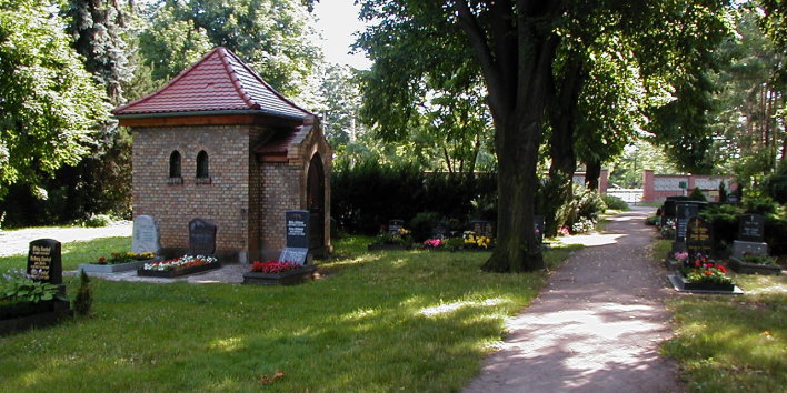 Zu sehen ist ein Friedhofsweg, an dessen Rande vereinzelte Gräber und eine Kapelle stehen
