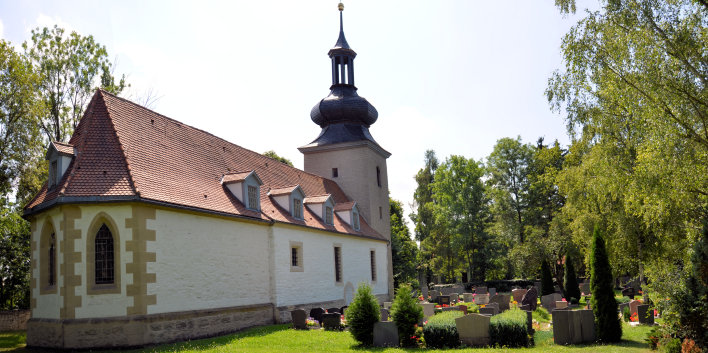 Neben ein paar Gräbern steht die hell verputzte Kirche mit Gauben auf dem mit Ziegeln gedeckten Dach. Der Chor wird von drei gothischen Fenstern bestimmt. Der aus natursteinen errichtete quadratische Turrm hat eine schiefergedeckte Kuppel mit Glockentürmchen als Abschluss.