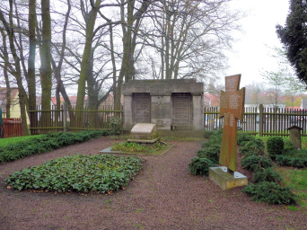 Monumentale Breitwandstele mit zwei vertieft liegenden Namensplatten, flankiert von zwei angedeuteten Säulen. In der Mitte ein Adlerrelief. Davor ein freistehender Pultstein und ein Holzkreuz mit weiteren Namen.