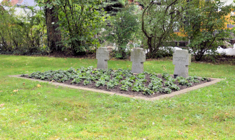 In einer Rasenfläche ist ein Rechteck mit einer Betonkante eingefasst und mit Stauden bepflanzt. In der Fläche stehen drei Granitkreuze mit den Namen der Opfer. Im Hintergrund stehen große Gehölze am Friedhofszaun.