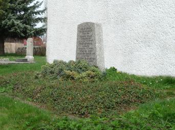 Vor einer Wand steht ein Granitgrabmal, welches sich nach oben leicht verjüngt.Auf dem Stein sind die Namen der toten eingraviert. Vor dem Stein steht ein kleiner Wachholder und flache Gehölze.