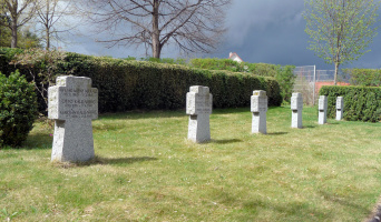 Eine Reihe Granitkreuze auf einer Wiese. Im Hintergrund bildet eine hohe Hecke den Abschluß der Fläche. Auf jedem Kreuz stehen drei Namen mit Lebensdaten.