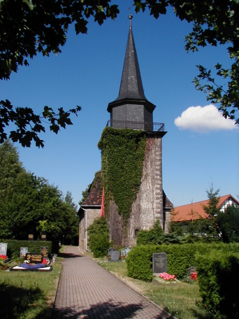 Blick entlang eines gepflasterten Friedhofsweges Richtung Kirche. Links und rechts liegen im Rasen einige Grabstätten, durch geschnittene Hecken getrennt. Im Hintergrund steht der mit Efeu bewachsene Kirchturm.