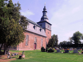 Blick über den Friedhof zur Kirche. Rechts befinden sich Grabstätten, die mit Blumen bepflanzt sind. Die kirche aus rotem Sandstein hat ein mit Schiefer gedecktes dach. Der quadratische Kirchturm aus gleichem Material hat eine verschieden gegliederte, mit Schiefer gedeckte Haube.