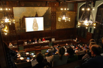 Blick in den Ratssitzungssaal von der Zuschauerempore. Im Saal viertelkreisförmige Stuhl-/Tischreihen, Präsidium und Rednerpult, Leinwand mit Gebärdendolmetscherin