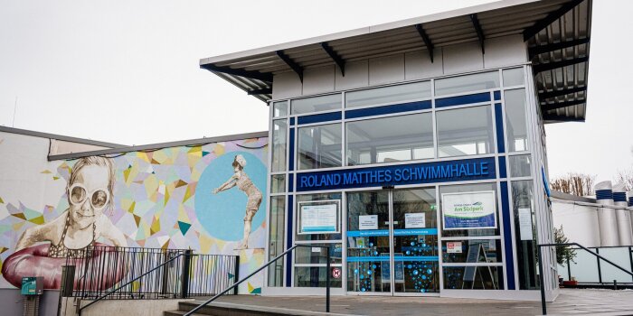 Der Haupteingang einer Schwimmhalle. Auf dem Schild steht Roland-Matthes-Schwimmhalle.