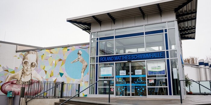 Der Haupteingang einer Schwimmhalle. Auf dem Schild steht Roland-Matthes-Schwimmhalle.