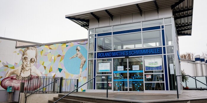 Der Haupteingang einer Schwimmhalle. Auf dem Schild steht Roland-Matthes-Schwimmhalle.