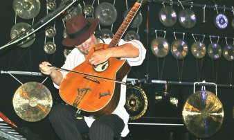 Foto: Peter-Kühnel © Peter Kühnel Ein Mann sitzt vor einer Reihe von Gongs an der Wand und streicht mit einem Bogen die Seite einer Gitarre.