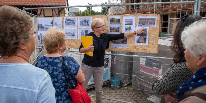 eine Frau zeigt auf verschiedene Fotos, die an einem Bauzaun hängen, mehrere Personen stehen vor ihr