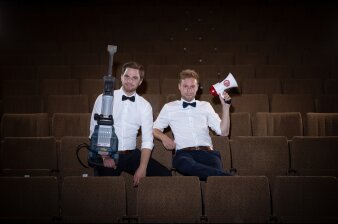 die beiden Künstler sitzen mit Presslufthammer und Megaphon im weißen Hemd und Fliege auf braunen Theatersesseln