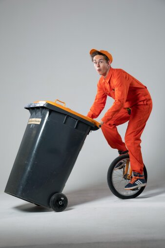 ein Mann im orangefarbenen Ganzkörperanzug fährt Einrad und schiebt dabei eine Mülltonne