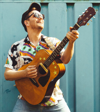 ein Mann mit buntem Hemd, Baseballkappe und Sonnenbrille spielt Gitarre