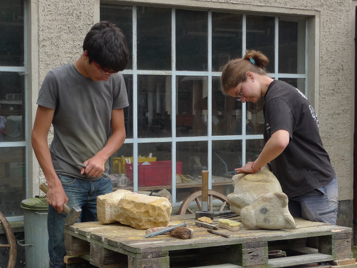 Zwei Jugendliche bearbeiten mit Hammer und Meißel zwei auf einem Tisch liegende Sandsteinblöcke.