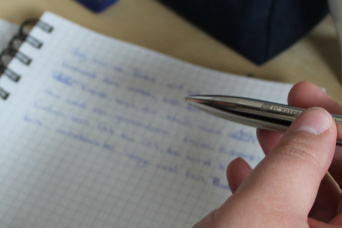 Schreibwerkstatt des Kulturrausch e. V. Eine Hand hält einen Füllfederhalter. Im Hintergrund ist ein mit Text beschriebenes Blatt zu sehen.