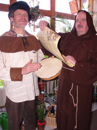 Zwei Männer in den historischen Kostümen von Bettelmönch und Spielmann. Der Spielmann hält ein Tamburin und der Mönch liest von einer Schriftrolle vor, die er in den Händen hält.