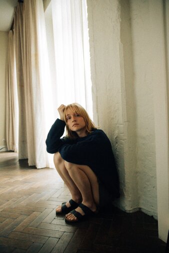 eine blonde Frau mit nackten Beinen und langem Pullover hockt an einer Wand auf dem Boden