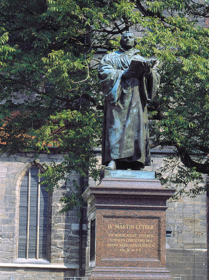 Erfurt, Lutherdenkmal vor der Kaufmannskirche Abbild einer Person auf einem Sockel, dahinter eine Kirche.