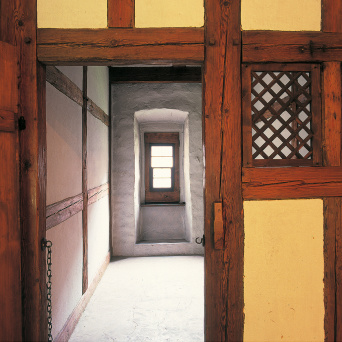 Im Vordergrund eine Fachwerkwand mit kleinem Gitterfenster und heruntergelassener Kette in der Türöffnung. Im Hintergrund kleiner Raum, weiß gestrichen, mit dicken Wänden.