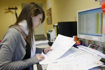 Eine Mitarbeiterin sitzt vor einem Computer. Vor ihr liegen Unterlagen, die Sie zur Bearbeitung des eingereichten Antrags benötigt.