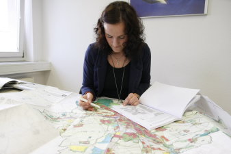 Eine Mitarbeiterin sitzt an einem Schreibtisch auf dem eine Stadtübersichtskarte ausgebreitet wurde und bearbeitet eingereichte Unterlagen zu einem Bauantrag.