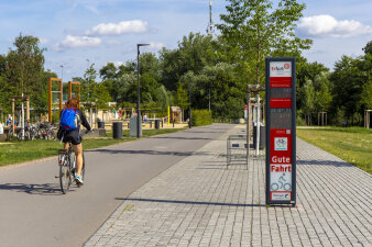 eine Radfahrerin fährt über einen Radweg, der durch einen Park verläuft, rechts eine Infostele mit Displays