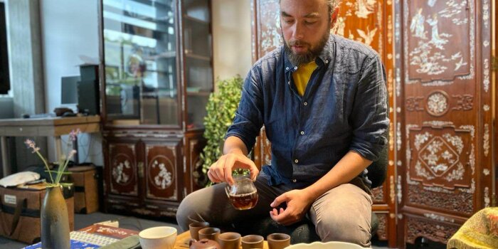 ein Mann sitzt vor einem umfangreichen Teeservice und hält ein Kännchen mit Tee in der Hand