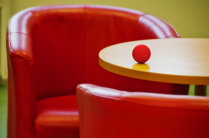 Das Foto zeigt zwei gegenüberstehende rote Sessel mit einem runden Tisch dazwischen, auf dem ein kleiner roter Ball liegt.