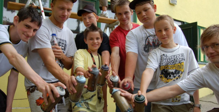 Eine Gruppe von sieben Jugendlichen und einem Sozialarbeiter stehen im Halbkreis und halten Spraydosen nach vorn gerichtet.