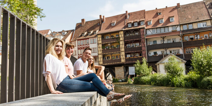 vier junge Menschen sitzen auf einer Mauer am Rande eines Flusses, im Hintergrund mehrere Fachwerkhäuser