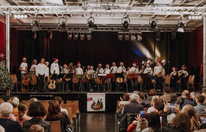 Spieler stehen mit ihren Gitarren und roten Weihnachtsmützen auf einer Bühne, im Vordergrund sitzen Gäste