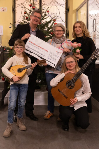 fünf Personen, davon halten zwei einen symbolischen Scheck, ein Kind hält eine Mandoline, eine Frau eine Gitarre