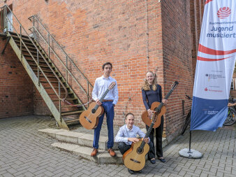 drei Jugendliche mit Gitarren neben einem Aufsteller mit Schriftzug "Jugend musiziert"
