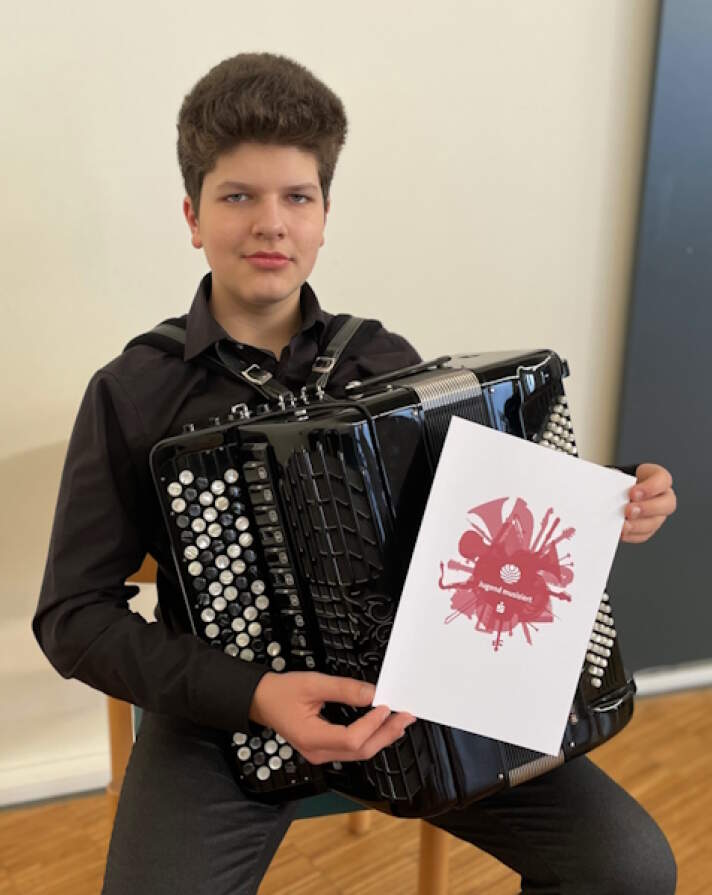 ein junger Musiker trägt ein Akkordeon und hält eine Jugend-musiziert-Urkunde in der Hand