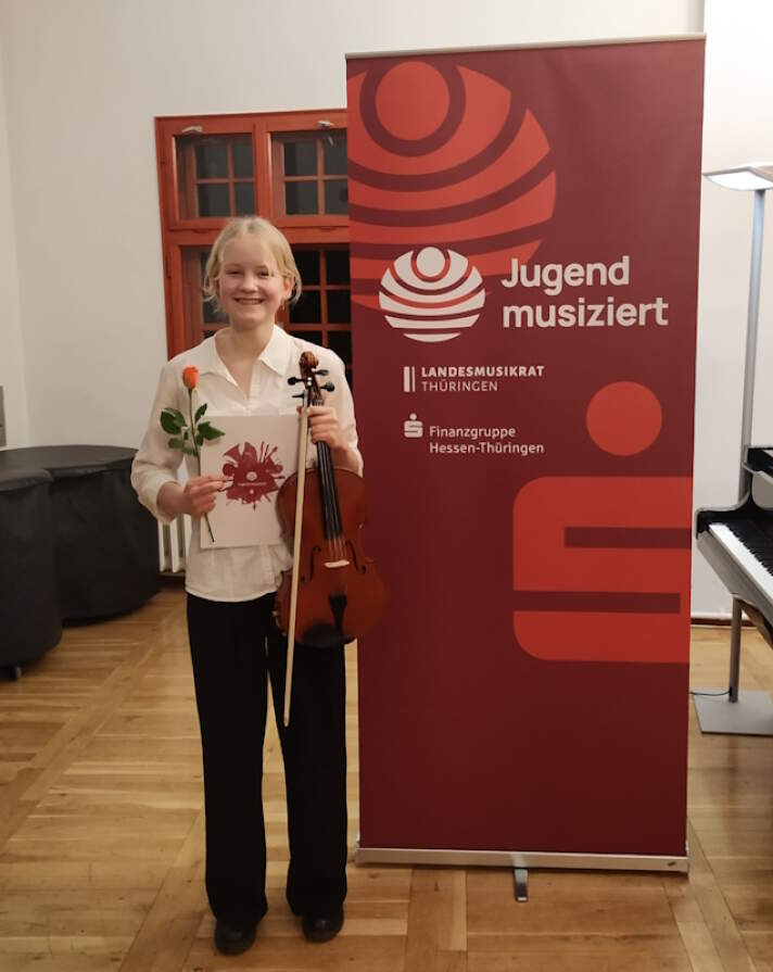 ein Mädchen steht neben einem Jugend-musiziert-Aufsteller und hält eine rote Rose, eine Urkunde und eine Violine