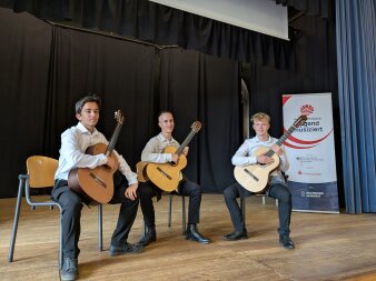drei Schüler mit Gitarren auf der Bühne mit Jugend musiziert Banner