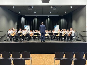 rund 25 junge Menschen sitzen in weißen Hemden bzw. Blusen und schwarzen Hosen auf einer Bühne und spielen Gitarre