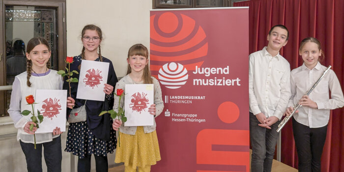 drei Kinder stehen neben einem Jugend-musiziert-Aufsteller, drei Mädchen halten eine rote Rose und eine Urkunde