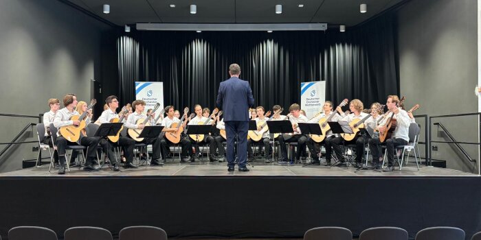 rund 25 junge Menschen sitzen in weißen Hemden bzw. Blusen und schwarzen Hosen auf einer Bühne und spielen Gitarre