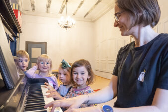 Kinder stehen am Klavier, während Lehrerin spielt