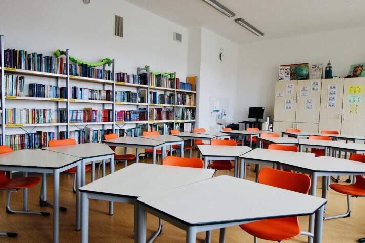 Das Foto zeigt viele Tische und Stühle in einem Raum sowie Bücherregale an der Wand.