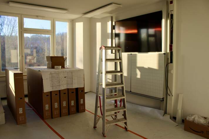 Das Foto zeigt ein Klassenzimmer mit Fernseher und Tafel an der Wand, Leiter und weiteren eingepackten Fernsehern.