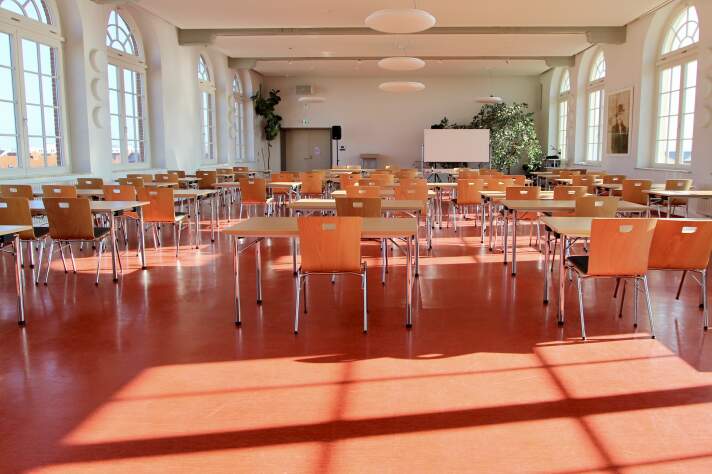 Das Foto zeigt die Aula der Schule mit einer Tafel und vielen Tischen und Stühlen.