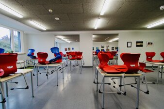 Das Foto zeigt die Mensa des GYM 10 mit roten Stühlen und vereinzelten blauen Stühlen.