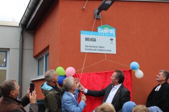 Das Foto zeigt drei Männer, die einen Vorhang vor dem Schild der Mensa zur Eröffnung herunterlassen